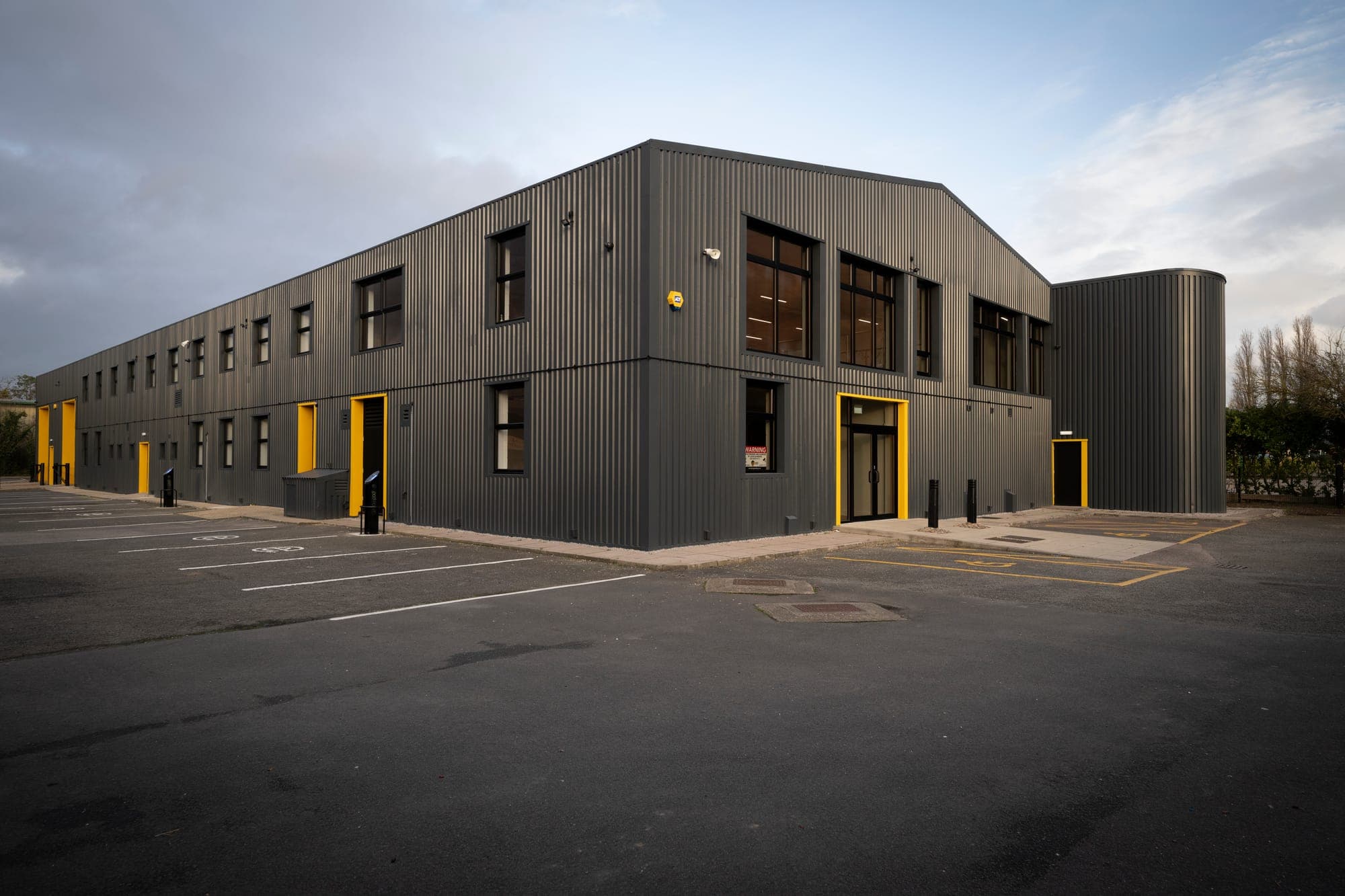 Novus: a two-storey industrial and office building in dark corrugated cladding with yellow door surrounds, seen across its car park at dusk.
